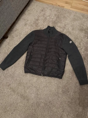 Mörkgrå cardiganjacka från Moncler - Snygg mörkgrå cardiganjacka från Moncler med quiltad front och stickade ärmar. Jackan har dragkedja framtill, två fickor med dragkedja och Moncler-logga på ärmen. Perfekt för lager-på-lager och en clean look. Priset kan diskuteras vid snabb affär.