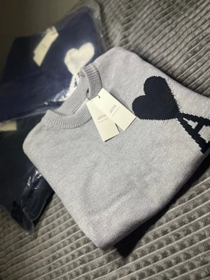Grå stickad tröja från Ami Paris - Snygg grå stickad tröja från Ami Paris med svart hjärta och A-logga på bröstet. Tröjan har rund halsringning och långa ärmar med ribbade muddar. Perfekt för en stilren och trendig look.