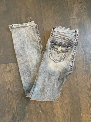 Grå tvättade jeans från True Religion - Säljer ett par grå tvättade jeans från True Religion med ikoniska sömmar och detaljerade bakfickor med knapp. Materialet är denim i bomull med en snygg sliten look. Knappt använda💞