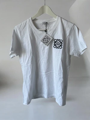 Vit Loewe t-shirt med svart logga - Säljer en clean vit t-shirt från Loewe med svart broderad logga på bröstet. Klassisk rund halsringning och korta ärmar. Tillverkad i mjuk bomull som känns skön mot huden. Perfekt för dig som gillar stilrena plagg med exklusiv känsla. Vet ej om äkta så släpper den billigt.