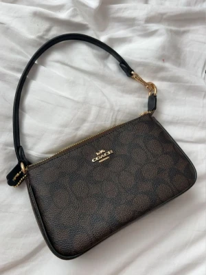 Coach Notila 19 - Säljer en stilren brun handväska från Coach med klassiskt monogrammönster och guldfärgade detaljer. Väskan har en avtagbar handledsrem och en diskret Coach-logga framtill. Materialet är en mix av canvas och läder. Digitalt kvitto från Coach ingår. Perfekt för dig som gillar tidlösa och eleganta accessoarer. Pris inte satt i sten kom jätte gärna med bud!!!