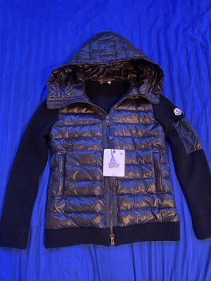 Svart dunjacka med huva från Moncler - Snygg svart dunjacka från Moncler med glansig framsida, stickade ärmar och rygg samt en stor huva. Jackan har dragkedja framtill, två fickor och Moncler-logga på ärmen. Perfekt för kalla vinterdagar och riktigt trendig look.