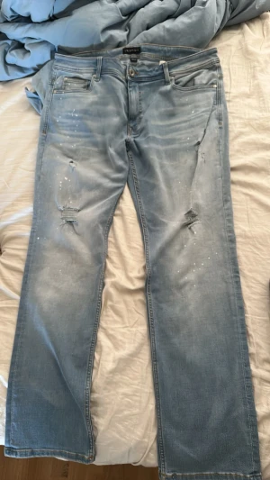 Ljusblåa jeans från Represent - Säljer ett par ljusblåa jeans med slitningar och små vita färgstänk på framsidan. Rätt stor i storleken 