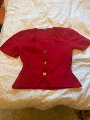 Röd blus med knappar från Gina Tricot - Snygg röd blus från Gina Tricot med figurnära passform och dekorativa guldknappar framtill. Blusen har korta ärmar och en v-ringad urringning.