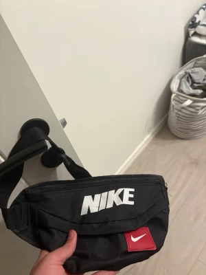 Svart magväska från Nike - Snygg svart magväska från Nike med stort vitt logotryck framtill och en röd detalj med vit swoosh. Justerbart band och dragkedja upptill. Perfekt för att bära det viktigaste när du är på språng.