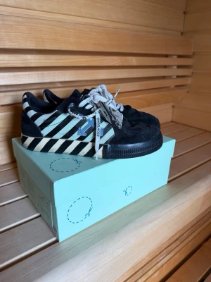 Off-White svarta och vita sneakers - Säljer ett par Off-White sneakers i svart mocka och textil med vita diagonalränder på sidan och Off-White-logga. Skorna har vita snören och en unik sula med svartvitt mönster. Snygg streetwear-look och ikoniska detaljer som zip-tie och pil-logga.