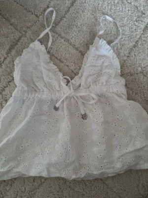 Vit broderad linnetopp från Censored - Söt vit linnetopp från Censored med tunna axelband, broderade blommor och dekorativ knytning framtill. Toppen har en luftig och lätt känsla, perfekt för varma dagar. Detaljerad med små hålbroderier och volangkant vid bysten.
