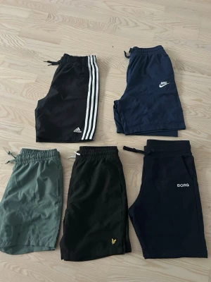 Mix av sportiga shorts från Adidas, Nike, Borg  - Fem par sportiga shorts i olika färger och märken: svarta Adidas med vita ränder, mörkblå Nike, gröna, svarta Lyle & Scott med gul logga och mörkblå Borg. Perfekta för träning eller chill. Alla har resårmidja och dragsko.