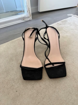 Svarta strappy pumps med smal klack från ASOS - Säljer ett par svarta pumps med tunna remmar och fyrkantig tå. Skorna har smal klack och justerbart spänne vid ankeln. Materialet ser ut att vara mocka eller mockaimitation och innersulan är ljusbeige. Perfekta för en trendig look. Endast använda en gång!