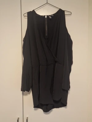 Svart omlott playsuit med lång ärm - Svart kort omlott playsuit med lång ärm och dragkedja i ryggen. playsuit  har en lös passform och är tillverkad i ett lätt, syntetiskt material. Perfekt för dig som gillar stilrena och enkla plagg med en twist.