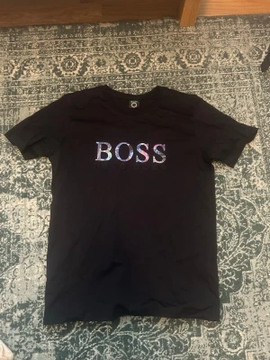 Hugo boss  - Storlek L men passar M också Svart t-shirt från Hugo Boss med stort, färgglatt BOSS-tryck på bröstet och diskret 'HUGO BOSS' under. Klassisk rund hals och korta ärmar. Perfekt för dig som vill sticka ut med en stilren men ändå unik look.