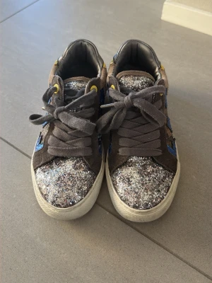 Glittriga sneakers med blå detaljer - Unika sneakers med grå bas, glittriga tår och coola blå och gula detaljer på sidorna. Skorna har breda grå skosnören och vit sula. Materialmix av textil och glitter ger en riktigt trendig look. Perfekta för dig som vill sticka ut i mängden.