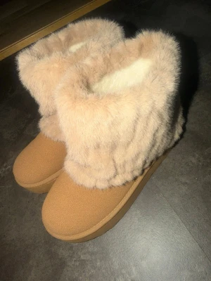 Beiga boots med fluffig fuskpäls - Mysiga tofflor i beige med hög, fluffig fuskpäls runt skaftet och mjuk insida. Yttermaterialet är mockaliknande och sulan är stabil. Perfekta för att hålla fötterna varma  under kyliga dagar.