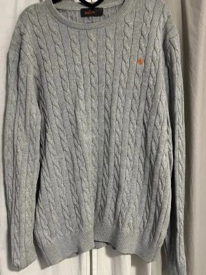 Ljusgrå kabelstickad tröja Morris - Kabelstickad ljusgrå tröja från Morris med rund halsringning och broderad orange logga på bröstet. Tröjan har långa ärmar och ribbade muddar vid ärmslut och nederkant. Perfekt för en stilren och klassisk look.