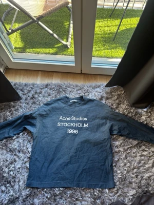 Mörkblå långärmad t-shirt Acne Studios -  långärmad t-shirt från Acne Studios med vit text 'STOCKHOLM 1996' på bröstet. Skön passform och klassisk rund halsringning. Perfekt för dig som gillar stilrena och trendiga plagg.