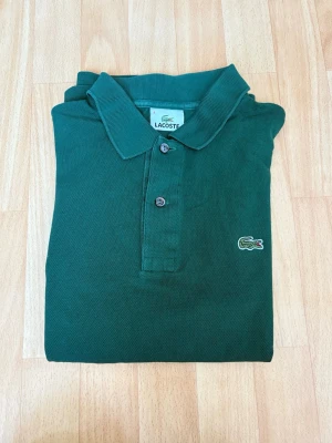 Grön piké t-shirt från Lacoste - Mörkgrön pikétröja från Lacoste med klassisk krage, korta ärmar och två knappar framtill. Storlek 4 (S).