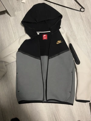 Nike hoodie i svart, grå och beige - Nike hoodie med dragkedja i svart, grå och beige färgblock. Guldigt Nike-märke på bröstet och praktisk ficka på ärmen. Tillverkad i mjukt material, perfekt för en chill och sportig look. Huva med dragsko och ribbade muddar för extra komfort.