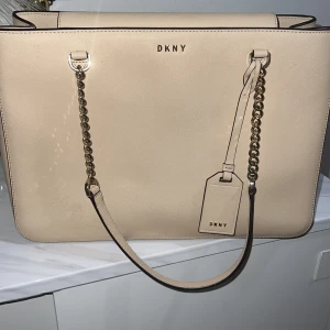 Beige axelväska från DKNY med kedjedetalj - Snygg beige axelväska från DKNY med stilren design och guldfärgade kedjedetaljer på axelremmen. Väskan är i slätt skinn och har en matchande tag med DKNY-logga framtill. Perfekt för dig som gillar klassiska och trendiga accessoarer. Priset kan disskuteras vid snabb affär!