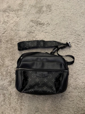 Louis Vuitton svart axelväska monogram - Snygg svart axelväska från Louis Vuitton med klassiskt monogrammönster i grått. Väskan har justerbar axelrem, en stor ficka med dragkedja framtill och är tillverkad i tåligt läder. Perfekt för dig som vill ha en stilren och lyxig accessoar.