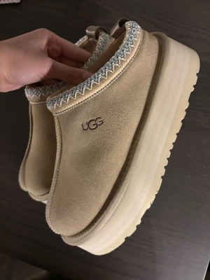 Beige UGG tofflor med platåsula - Snygga beige tofflor från UGG med hög platåsula och broderad kant i vitt och blått. Utsidan är i mocka och insidan är fodrad med mjuk ull för extra komfort. Perfekta för att hålla fötterna varma och stilrena hemma.
