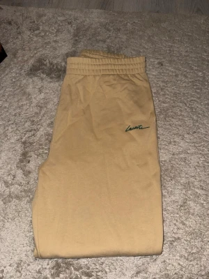 Beige mjukisbyxor från Lacoste - Säljer ett par beige mjukisbyxor från Lacoste med elastisk midja och grön Lacoste-logga framtill. 