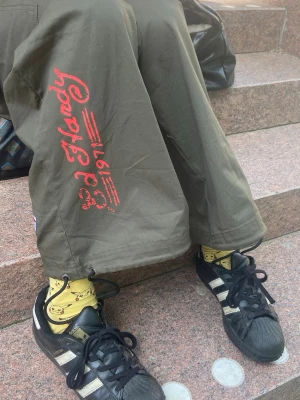 Gröna cargobyxor från Ed Hardy - Säljer ett par gröna cargobyxor från Ed Hardy med bred passform och röda tryckdetaljer på benet. Byxorna har stora sidofickor och cool streetwear-vibe. Perfekta för dig som gillar en avslappnad och trendig stil.