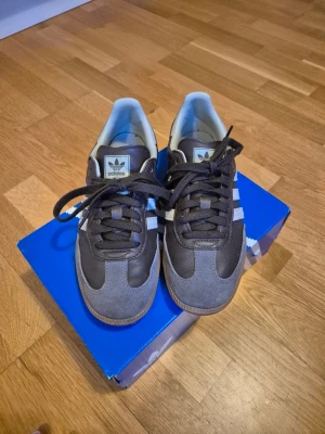 Adidas Samba sneakers brun/grå 39 1/3 - Snygga Adidas Samba sneakers i brunt och grått med klassiska vita ränder på sidorna. Ovandelen är i läder och mocka, med gummisula och brunt snörning. Perfekta för dig som gillar retrostil och streetwear.