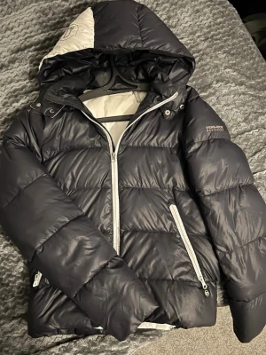 Svart pufferjacka från Penn-Rich - Snygg svart pufferjacka från Penn-Rich med vit dragkedja och detaljer. Jackan har huva med vit insida och två dragkedjeförsedda fickor. Perfekt för kalla vinterdagar och riktigt skön att bära. Klassisk quiltad design och varm vaddering.