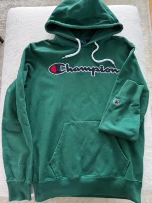 Grön Champion hoodie med broderad logo - Snygg grön hoodie från Champion med stor broderad logga över bröstet och klassisk känguruficka. Huva med vita dragsnören och liten Champion-logga på ärmen. Mjuk bomullsblandning som är perfekt för chill eller streetwear.