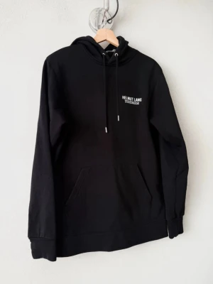 Svart hoodie från Helmut Lang - Svart hoodie från Helmut Lang med vit logga tryckt på bröstet. Klassisk modell med huva och dragsnören, samt en stor magficka framtill. Tillverkad i mjukt bomullsmaterial och har ribbade muddar vid ärmslut och nederkant.