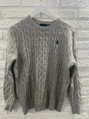 Grå kabelstickad tröja från Polo Ralph Lauren - Kabelstickad grå tröja från Polo Ralph Lauren med klassisk rund halsringning och svart broderad logotyp på bröstet. Tröjan har långa ärmar och ribbade muddar vid ärmslut och nederkant. Perfekt för lager-på-lager och stilren look.