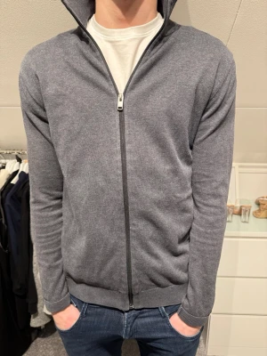 Grå ziptröja med hög krage - Snygg grå ziptröja med hög krage och hel dragkedja framtill. Tröjan är stickad i ett mjukt material och har långärmad design med ribbade muddar vid ärmslut och nederkant. Perfekt att slänga på sig över en t-shirt för en clean look.
