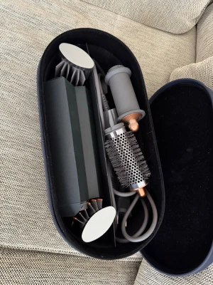 Dyson Airwrap multistyler med tillbehör - Säljer en Dyson Airwrap multistyler i snygg grå och kopparfärg. Setet innehåller flera olika borsthuvuden, locktillbehör och en rundborste, allt i en elegant oval förvaringsväska. Perfekt för dig som vill styla håret på olika sätt med ett enda verktyg.