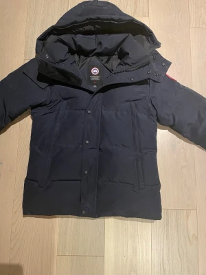 Canada Goose wyndham - Mörkblå Canada Goose med huva och diskret logga på ärmen. Jackan har dragkedja och knappar framtill samt två stora fickor. Perfekt för kalla vinterdagar. Pälsen är hög kvalitet och går att ta av. 