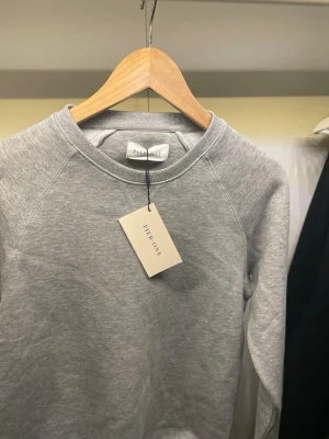Grå sweatshirt från Pier One - En stilren gråmelerad sweatshirt från Pier One med rund halsringning och raglanärmar. Tröjan är tillverkad i mjukt material och passar perfekt för en avslappnad och trendig look. Enkel design utan tryck eller detaljer, vilket gör den lätt att matcha.
