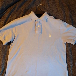Ljusblå pikétröja från Ralph Lauren - Ljusblå pikétröja från Ralph Lauren. Vita loggan broderad på bröstet. Tröjan är i mjuk bomull och har korta ärmar.
