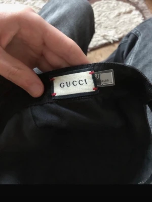 Svart keps från Gucci med monogram - Snygg svart keps från Gucci med diskret GG-monogram över hela skärmen. Klassisk form med böjd skärm och justerbar passform. Tillverkad i slitstarkt tyg som känns exklusivt och trendigt. Perfekt accessoar för att lyfta din outfit.