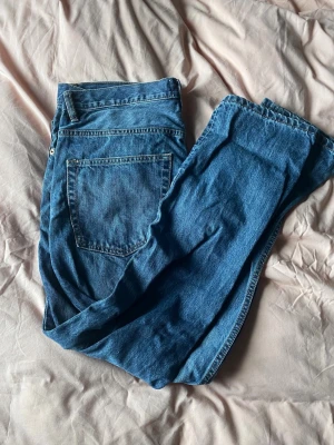 Blå raka jeans i klassisk modell - Säljer ett par blå jeans med rak passform och fem fickor. Jeansen har normal midja och är tillverkade i ett klassiskt denimtyg. Perfekta för en avslappnad och tidlös stil.