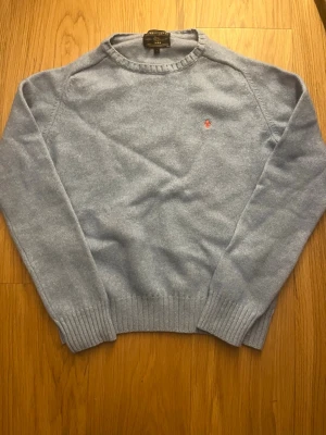 Ljusblå stickad Morris tröja  - Ljusblå stickad tröja från morris med rund halsringning och diskret röd logga broderad på bröstet. Tröjan har långa ärmar och ribbade muddar vid ärmslut och nederkant. Perfekt för lager-på-lager och enkel att matcha.