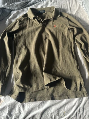 Olivgrön långärmad skjorta Polo Ralph Lauren - Klassisk olivgrön långärmad skjorta från Polo Ralph Lauren med krage och liten röd broderad logga på bröstet. Tillverkad i mjuk bomull och har knappar vid halsen. Perfekt för en avslappnad men ändå stilren look. XS men lappen inuti är borta