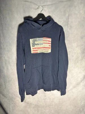 hoodie från Ralph Lauren - Mörkblå hoodie från Ralph Lauren med tryckt amerikansk flagga och text på bröstet. Klassisk känguruficka framtill och huva. Mjuk bomullskvalitet och avslappnad passform, perfekt för en chill stil. Små defekter. Storlek L sitter mer som M
