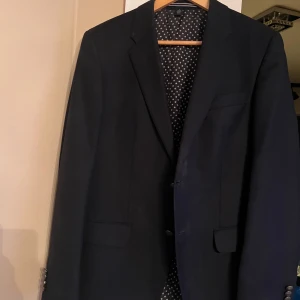 Mörkblå kavaj från Tommy Hilfiger - Stilren mörkblå kavaj från Tommy Hilfiger med klassisk passform. Kavajen har två framfickor med lock, smala slag och fyra dekorativa knappar vid ärmsluten med ingraverad logga. Perfekt för en dressad look och tillverkad i ett tyg med struktur.