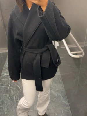 Svart omlottkappa med knytskärp - Stilren svart kappa i omlottmodell med brett slag och knytskärp i midjan. Kappan har en rak passform och är tillverkad i ett mjukt tyg som ger en snygg siluett. Perfekt för dig som gillar minimalistisk och trendig stil.