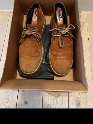 Bruna mockakängor med snörning - Säljer ett par klassiska bruna kängor i mocka med snörning och kontrastsömmar runt sulan. Skorna har en enkel och stilren design som passar till många olika outfits. Perfekta för dig som gillar en avslappnad men ändå snygg look.