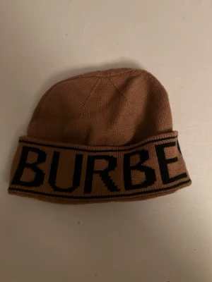 Beige Burberry mössa med logga - Säljer en beige stickad mössa från Burberry med bred vikt kant och stor svart logotyp runt om. Mössan är i mjukt material och har en klassisk, avslappnad passform som passar perfekt till höst och vinter.
