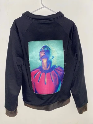 Svart hoodie med konstnärligt tryck - Svart hoodie med dragkedja och två fickor framtill. På ryggen finns ett stort, färgstarkt konstnärligt tryck av en person i neonfärger och röd tröja. Hoodien har muddar vid ärmslut och nederkant samt en liten broderad logga på bröstet.