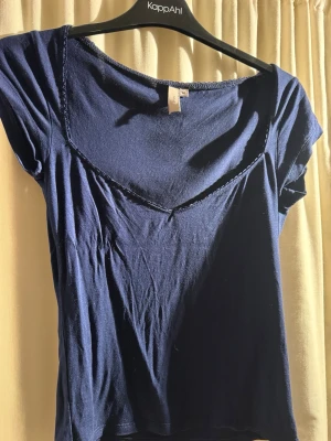 Mörkblå topp med spetskant från Nelly - Snygg mörkblå topp från Nelly med korta ärmar och v-ringad halsringning dekorerad med en fin kant. Sparsamt använd, säljer då den inte passar