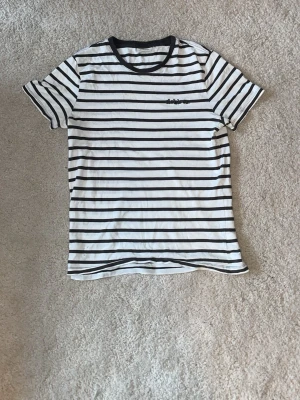 Randig t-shirt från Dobber - Snygg vit t-shirt med svarta horisontella ränder från Dobber. T-shirten har svart kant vid halsen och broderad logga på bröstet. Perfekt för en avslappnad stil och enkel att matcha med jeans eller shorts.