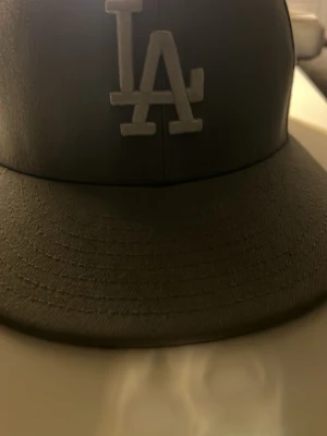 Svart LA-keps från New Era - Säljer en svart keps från New Era med LA-logga broderad framtill i vitt. Klassisk flat brim och MLB-logga baktill. Kepsen är gjord i ett material som liknar konstläder och har en modern streetwear-look.