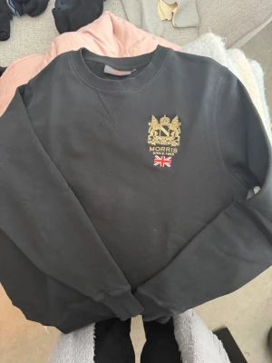 Svart Morris sweatshirt med brodyr - Svart sweatshirt från Morris med guldbroderad logga och brittisk flagga på bröstet. Klassisk rund halsringning och ribbade muddar. Perfekt för en stilren och avslappnad look.